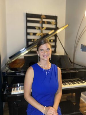 Brittany is smiling and sitting at her grand piano, wearing a blue dress and a necklace, and this photo was taken just before a live performance online.